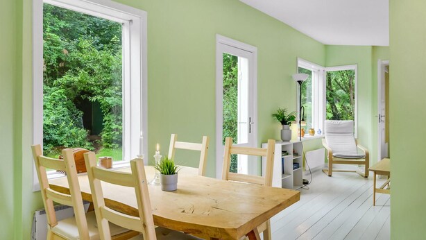 A room with a wooden dining table and chairs, large windows with a view of greenery, and a white ceiling.
