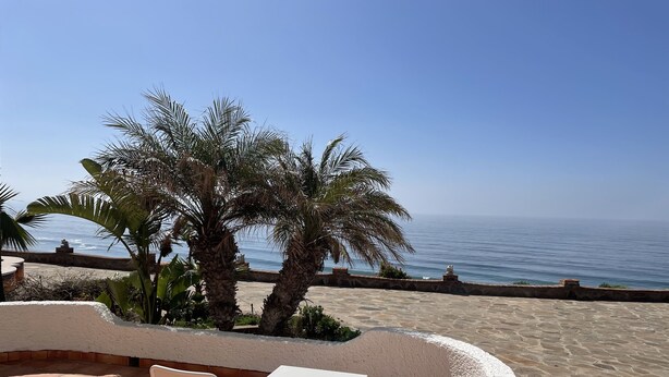 Une terrasse extérieure avec deux chaises en métal blanc et une table, donnant sur une plage de sable et la mer.