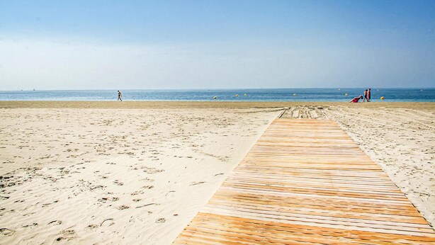 Una playa con un paseo marítimo de madera que lleva al agua, arena y un cielo despejado.
