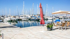 Un puerto deportivo con numerosos veleros amarrados, una sombrilla roja y mobiliario exterior con sillas y una maceta.