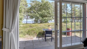 Ein Zimmer mit einer Schiebeglastür, die auf eine Terrasse mit Tisch und Stühlen führt. Blick auf Bäume und ein Gewässer.