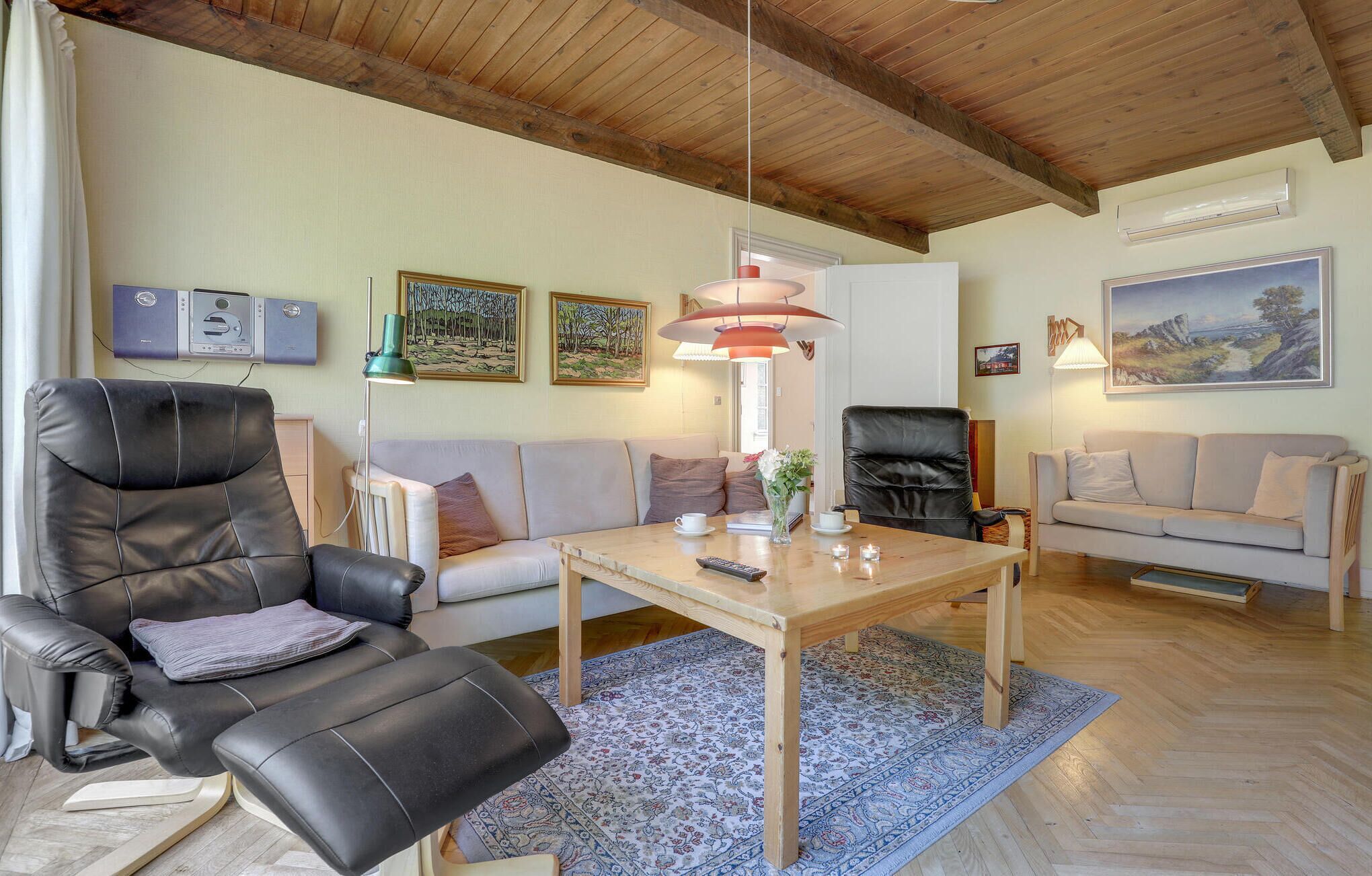 A living room with a wooden ceiling, a sofa, a black leather chair, a wooden coffee table, and a patterned rug.