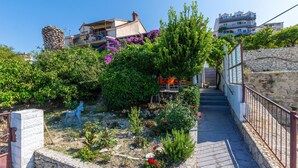 Ein schmaler Steinweg führt zu einem Gebäude mit Balkon, umgeben von üppiger Vegetation und blühenden Pflanzen.