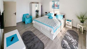 Ein modernes Schlafzimmer mit Bett, Sessel, kleinem Tisch, Nachttisch, Topfpflanze und einem Bild an der Wand.