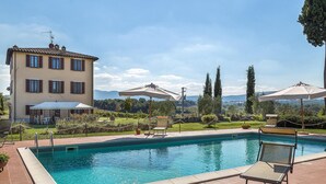 A swimming pool with lounge chairs and umbrellas, surrounded by a landscaped garden and a building with multiple windows. - Stunning home in Chiusi (Chiusi)