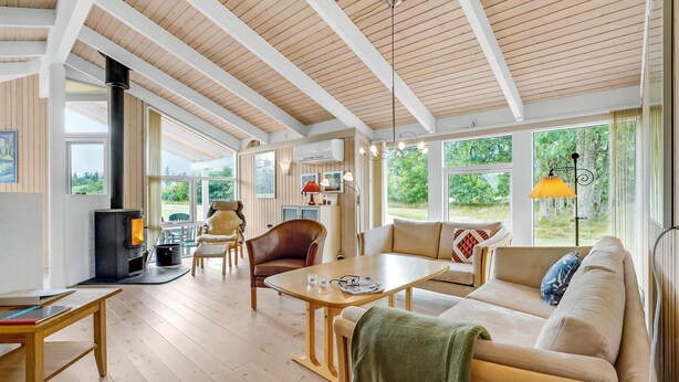A cozy, well-lit room with a wooden ceiling, a fireplace, a sofa, a table, and large windows offering a view of greenery outside.