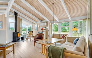 Une pièce chaleureuse et bien éclairée, avec un plafond en bois, une cheminée, un canapé, une table et de grandes fenêtres donnant sur un paysage verdoyant.