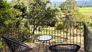 Een terras met twee stoelen en een tafel, omringd door een metalen hek, met uitzicht op een landelijk landschap met bomen en heuvels.