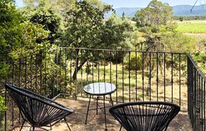 A patio with two chairs and a table, surrounded by a metal fence, overlooking a rural landscape with trees and hills.