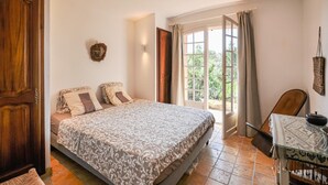 Een slaapkamer met een bed, een houten deur, een raam met gordijnen, en een klein tafeltje met een stoel.