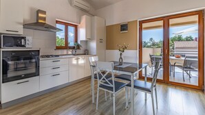 A modern kitchen with white cabinets, a built-in microwave, a stove, and a large window. There is a dining area with a table and chairs, and a sliding glass door leading to an outdoor patio. - Lovely home in Barbariga (Barbariga)
