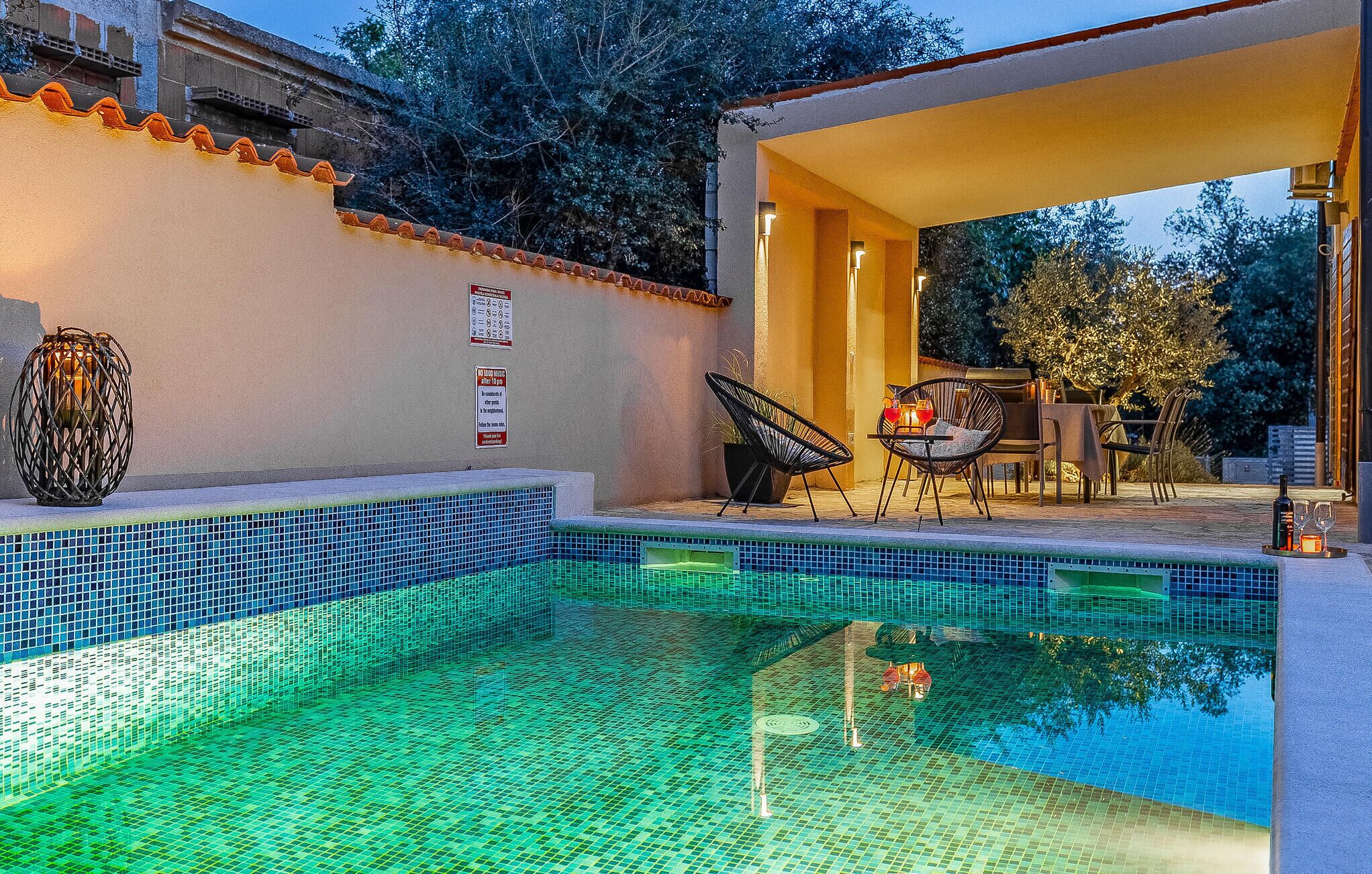 Una piscina con borde de azulejos, un área de descanso con mobiliario exterior y una estructura de pérgola.