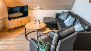 A modern living room with a flat-screen TV, a round wooden coffee table, a black leather sofa with blue cushions, and a small potted plant. - Cozy home in Gråsten with kitchen (Gråsten)