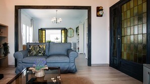 Ein Wohnzimmer mit einer Couch, einem Couchtisch mit Blumen in einer Vase und Blick in die Küche durch eine Glastür.
