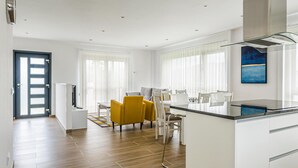 A modern kitchen with a dining area, featuring a central island, white cabinetry, and a large window with blinds. - Gorgeous home in Noia (Noia)