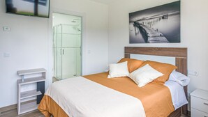 A hotel room with a bed, a glass shower enclosure, a TV mounted on the wall, and a painting of a pier. - Gorgeous home in Noia (Noia)