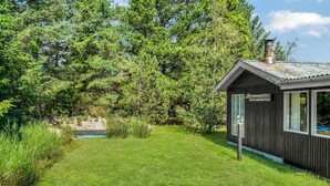 Une petite cabane en bois avec une cheminée, entourée d’une végétation luxuriante et sous un ciel bleu clair.