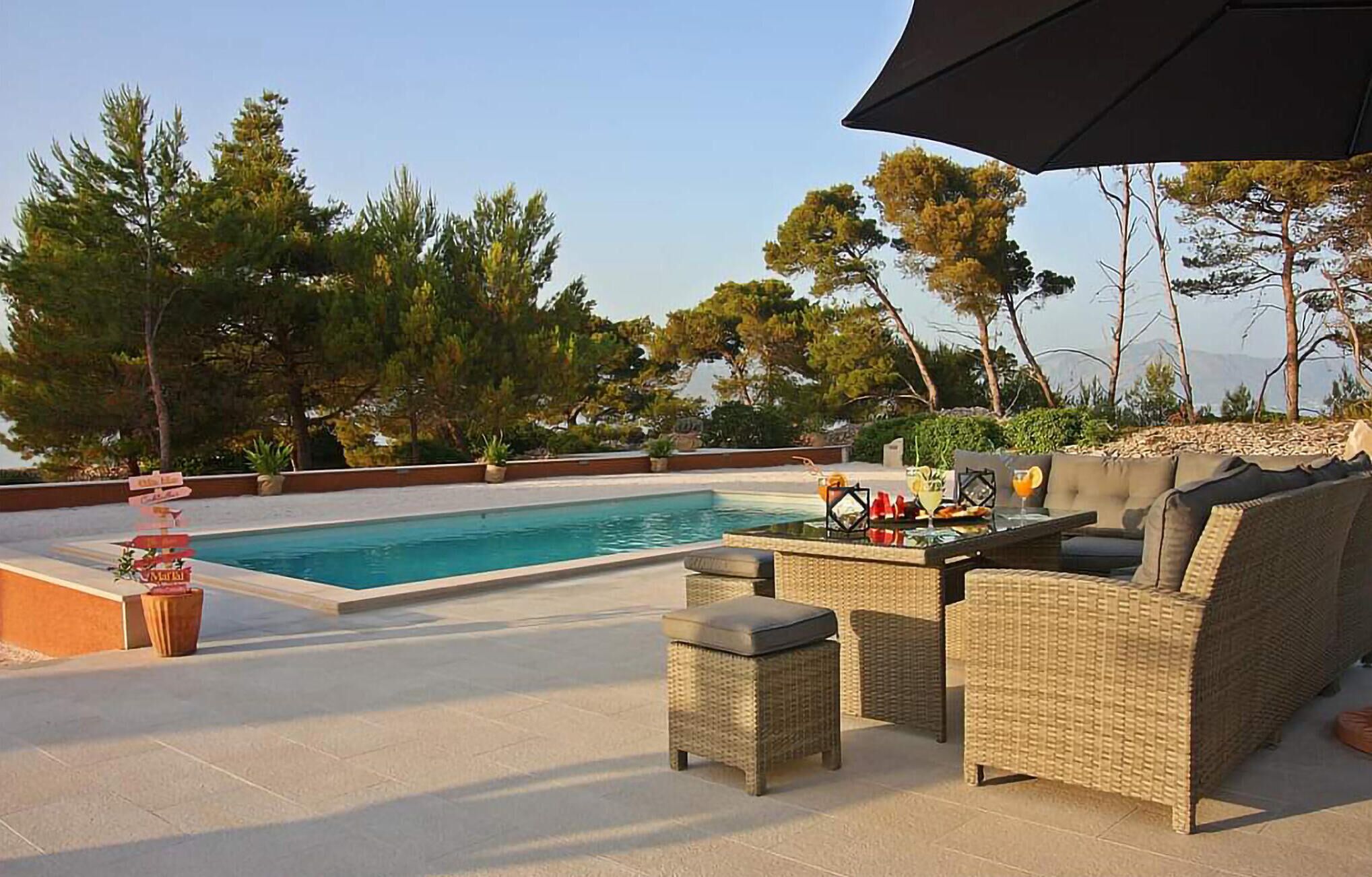 Uma área de piscina com um sofá e mesa de vime, cercada por árvores e um céu limpo.