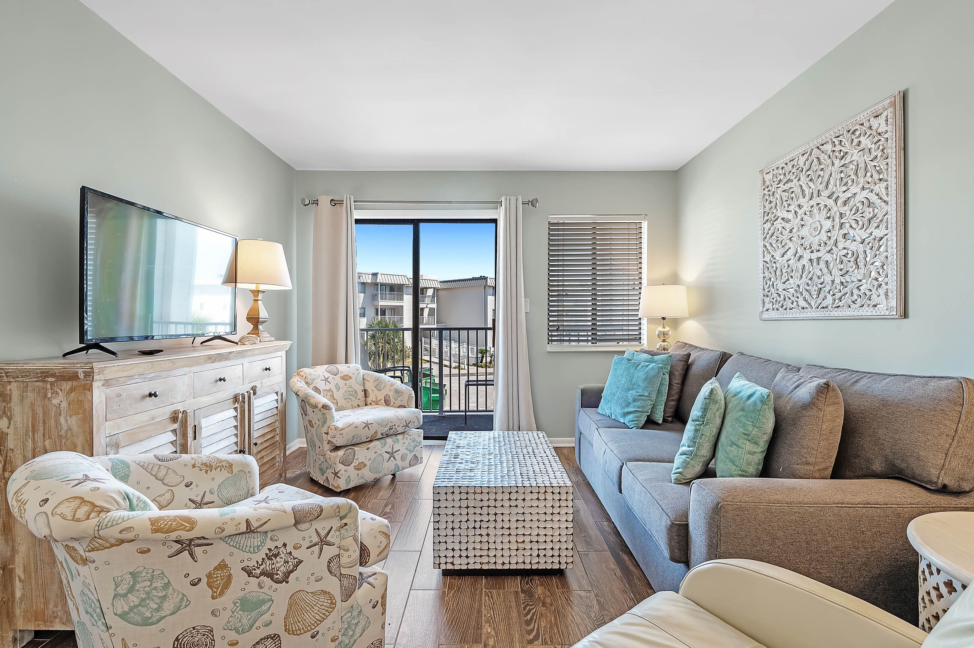 A living room with a sofa, armchairs, a coffee table, a TV, and a balcony with curtains.