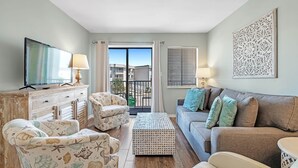 Un salon comprenant un canapé, des fauteuils, une table basse, un téléviseur et un balcon avec des rideaux.