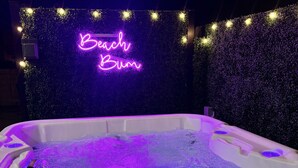 Ein Whirlpool mit violetter Beleuchtung, umgeben von einer mit Grünpflanzen verzierten Wand und einem Neonlichtschild mit dem Schriftzug 'Beach Bum'.