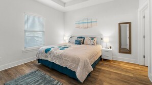 Ein Schlafzimmer mit Bett, Nachttischen, Deckenventilator, Fenster mit Jalousien, Wandspiegel und Teppich.