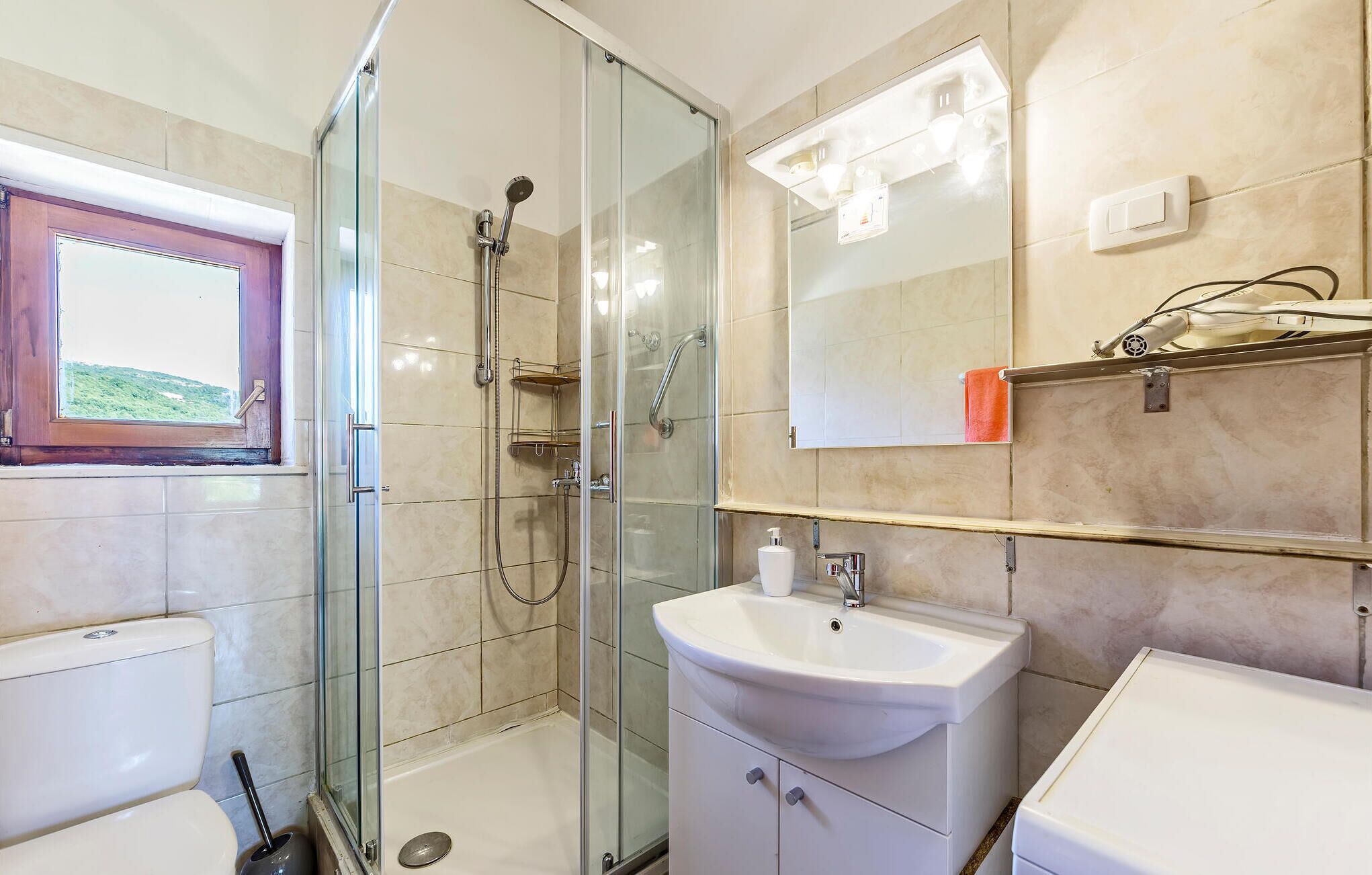 Ein Badezimmer mit Duschkabine, Toilette, Waschbecken und einem Fenster mit Blick ins Grüne.