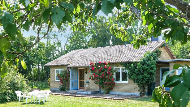 Ein kleines Haus mit Ziegeldach, einer Haustür und einem kleinen Vordach. Vor dem Haus befindet sich eine Rasenfläche mit ein paar Stühlen und einem Tisch.
