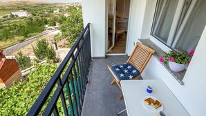 Un balcon donnant sur un paysage rural, une chaise en bois avec un coussin, une petite table avec une assiette et une tasse, et une plante en pot.