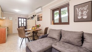 Ein modernes Wohnzimmer mit grauem Sofa, einer Essecke mit Tisch und Stühlen, einer Küche mit Herd und einem Fenster mit Blick auf Bäume.