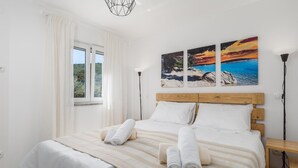 A bedroom with a large bed, white bedding, a view of the outdoors through a window, and three paintings on the wall.