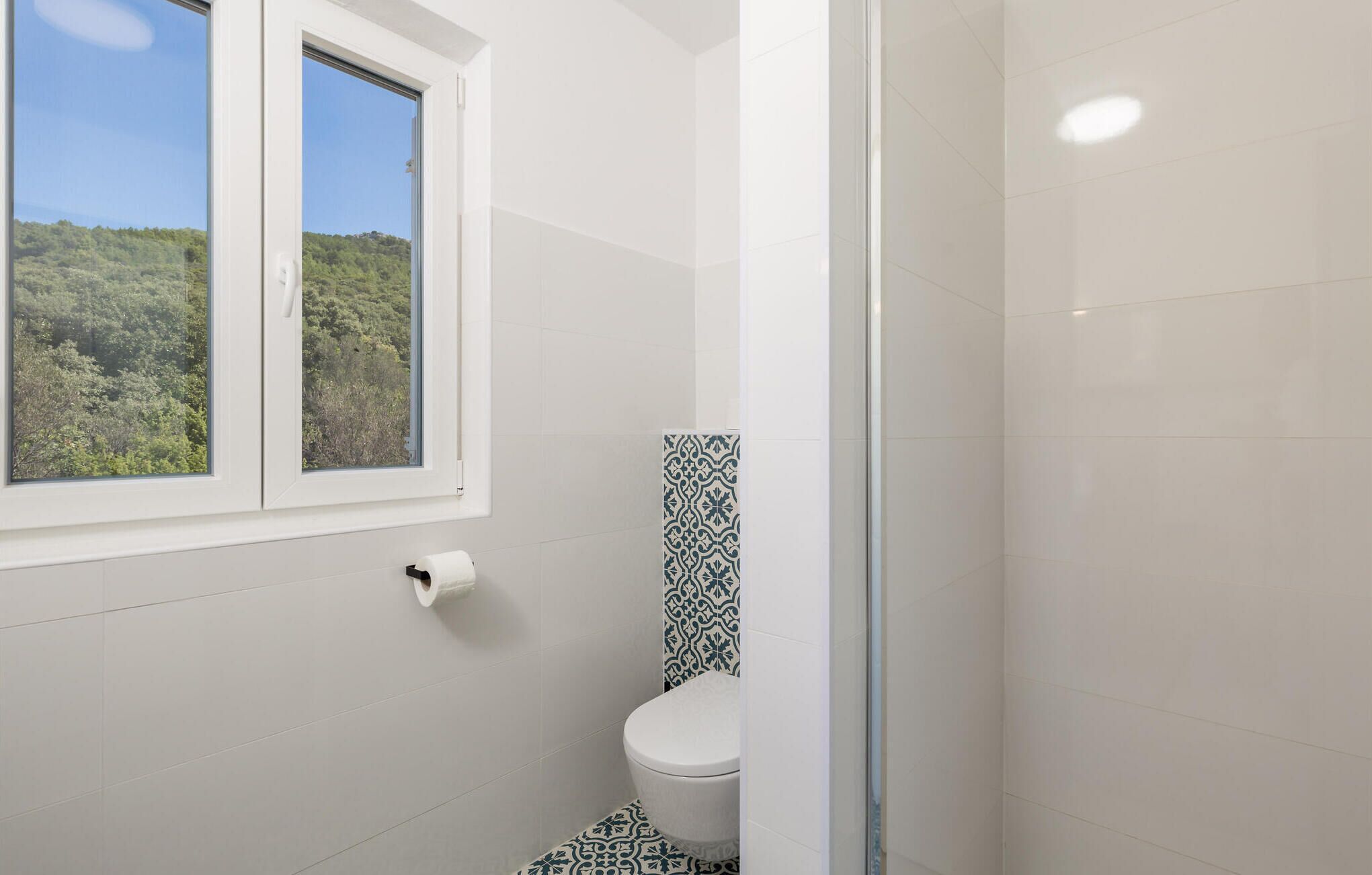 Une salle de bain avec des toilettes, une fenêtre donnant sur un espace verdoyant et un mur recouvert de carreaux décoratifs.