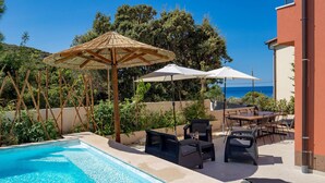 A poolside area with a pool, outdoor furniture, umbrellas, and a view of the sea.