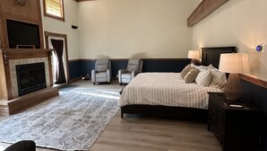 Un dormitorio amplio con una cama grande, chimenea, dos sillones, ventilador de techo y una mesita de noche con lámpara.