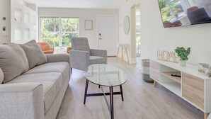 A modern living room with a sofa, armchair, and coffee table. There is a TV mounted on the wall, a bookshelf, and a door leading to another room.