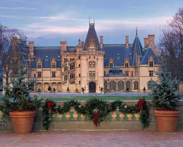 A large, historic building with a central tower and multiple wings, surrounded by a brick pathway and potted plants.