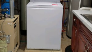Een compacte, witte wasmachine in een technische ruimte met zichtbare leidingen en een boiler.