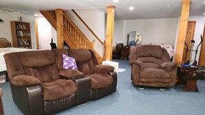 Een woonkamer met een bruine leren bank, een bruine fauteuil, een houten trap en een boekenkast.