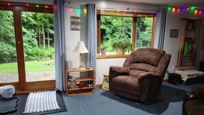 Een woonkamer met een bruine fauteuil, een klein houten schapje, een lamp en uitzicht op groen door grote ramen.