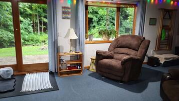 A living room with a brown recliner, a small wooden shelf, a lamp, and a view of greenery through large windows.