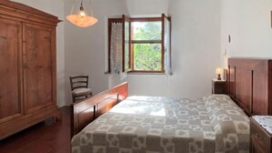 A bedroom with a wooden wardrobe, a bed with a patterned bedspread, a chair, a nightstand with a lamp, and a window with curtains. - Apartment Sonia by Interhome (Riparbella)