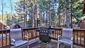 Eine Holzterrasse mit Grill und zwei Stühlen, umgeben von hohen Bäumen.
