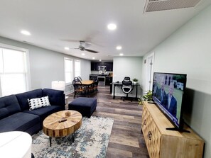 A modern living room with a dark blue sofa, a round wooden coffee table, a TV on a wooden cabinet, and a kitchen in the background. - `Charlie`s by the Bay` # 4 (New Baltimore)
