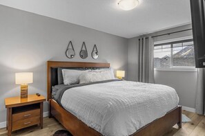 A bedroom with a wooden bed, bedside lamps, a nightstand, and a window with a view. - Rocky Mountain Condo, Grotto Mtn Views, Hot Tub (Dead Man's Flats)