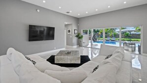 A modern living room with a white sofa, a coffee table, and a large TV on the wall. - Luxury Unique Property 4-3 - Entire House - Heated Pool - Lg Yard (West Palm Beach)