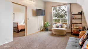 Una sala de estar acogedora con un sofá, una silla, un televisor y una estantería. Se ve una ventana con cortinas y una planta en el suelo.