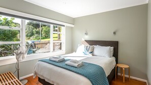 Ein Schlafzimmer mit großem Fenster, einem Bett mit blauer Steppdecke, einem Nachttisch und einem Standventilator.