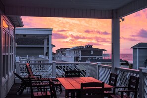 A covered outdoor seating area with tables and chairs, overlooking a residential neighborhood and the ocean during sunset. - Beautifully Decorated, 3 King Master Bedrooms, AND a Heated Pool by the Ocean (Ocean Isle Beach)