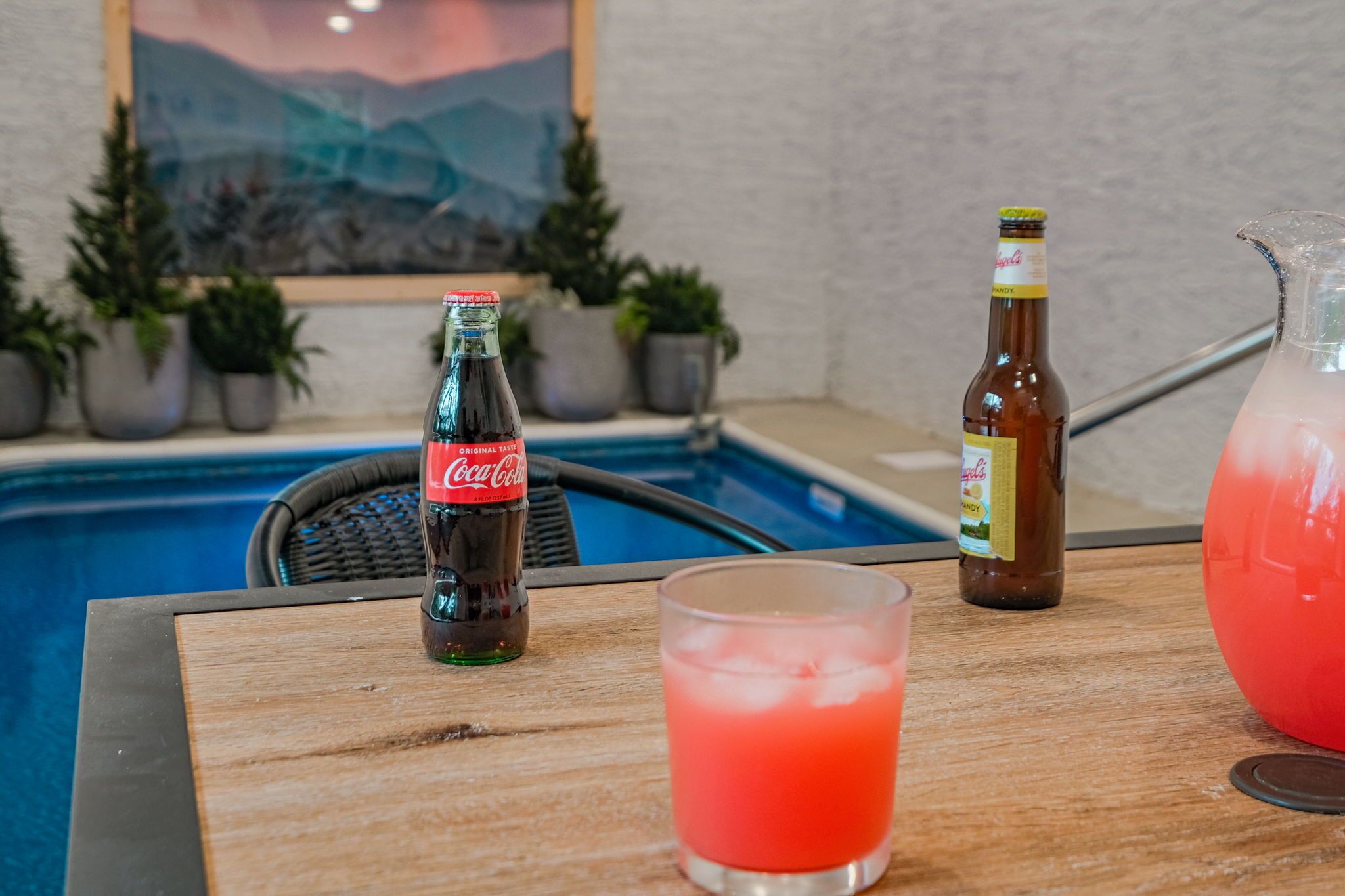 A poolside table with a Coca-Cola bottle, a pitcher of pink drink, and a bottle of beer.