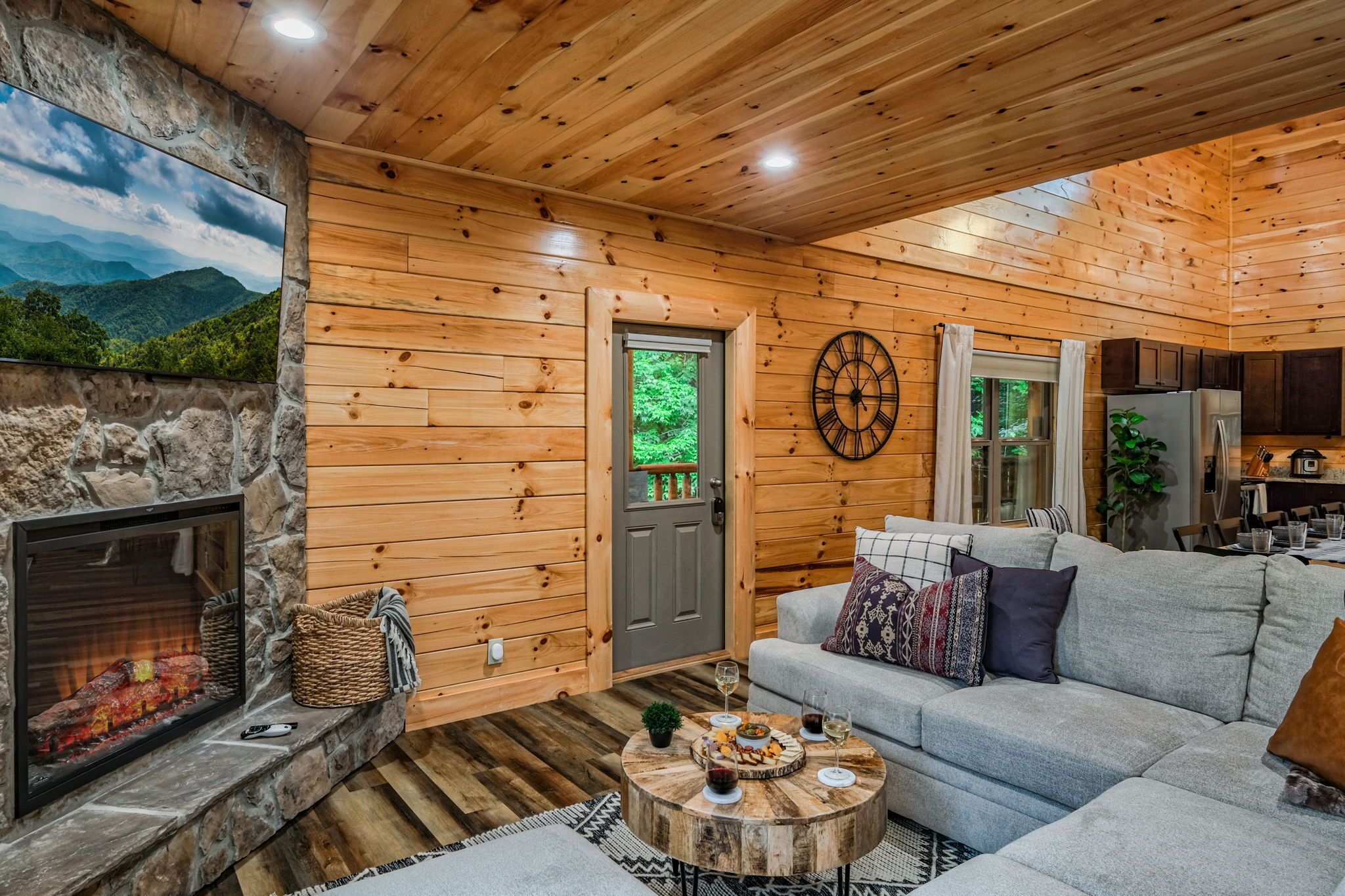 A cozy cabin interior with a stone fireplace, wooden walls, a gray sofa, and a dining area.